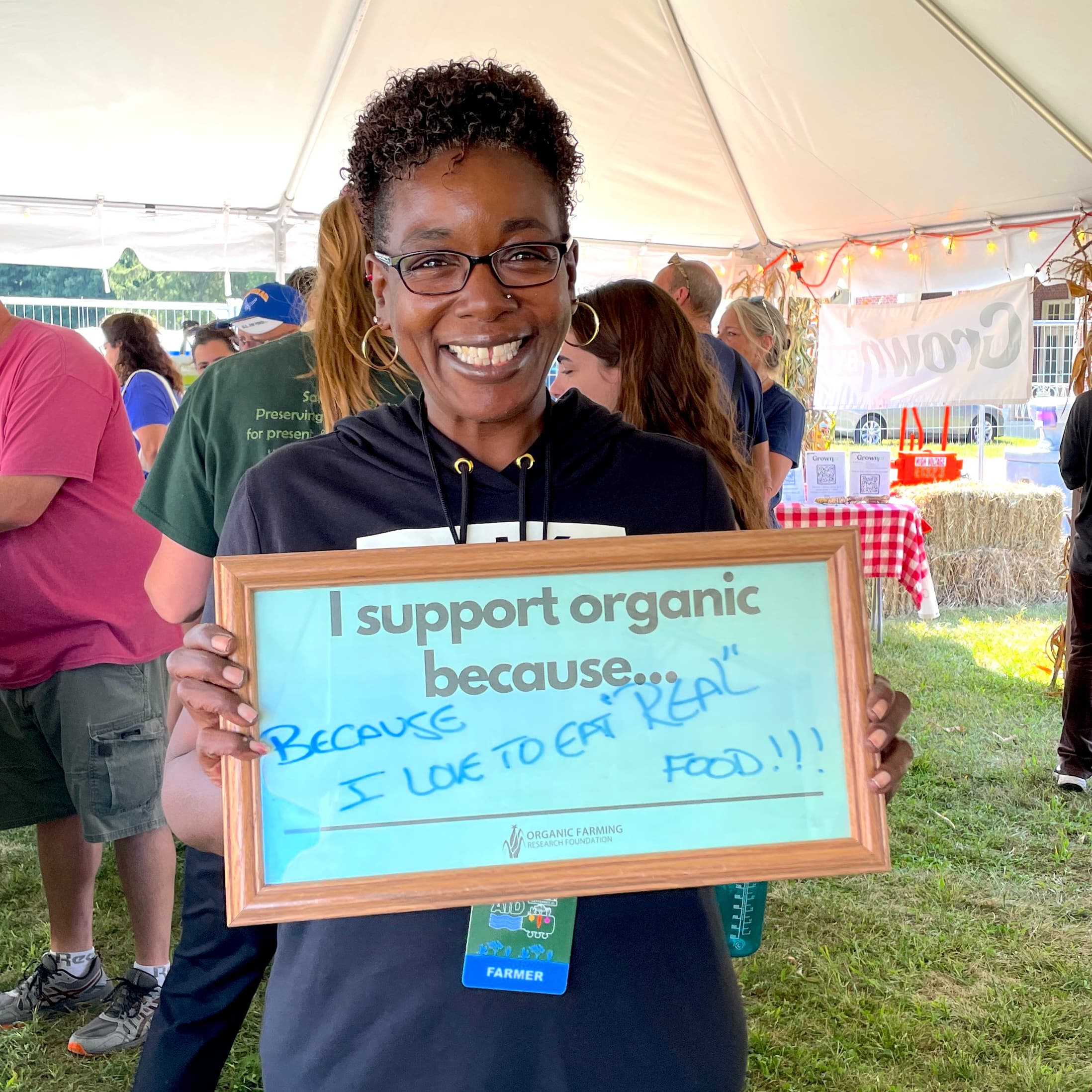 Farm Aid Organic Supporter Image farmer with glasses holding a sign that reads "I support organic because" and she wrote on the blank line "i love to eat 'real' food"